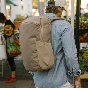 Person wearing a CIVIC Half Zip 22L in Fossil Brown backpack in an outdoor market with people and plants.