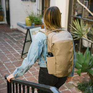 Person wearing a CIVIC Half Zip 22L in Fossil Brown backpack in an outdoor setting with plants and a building in the background