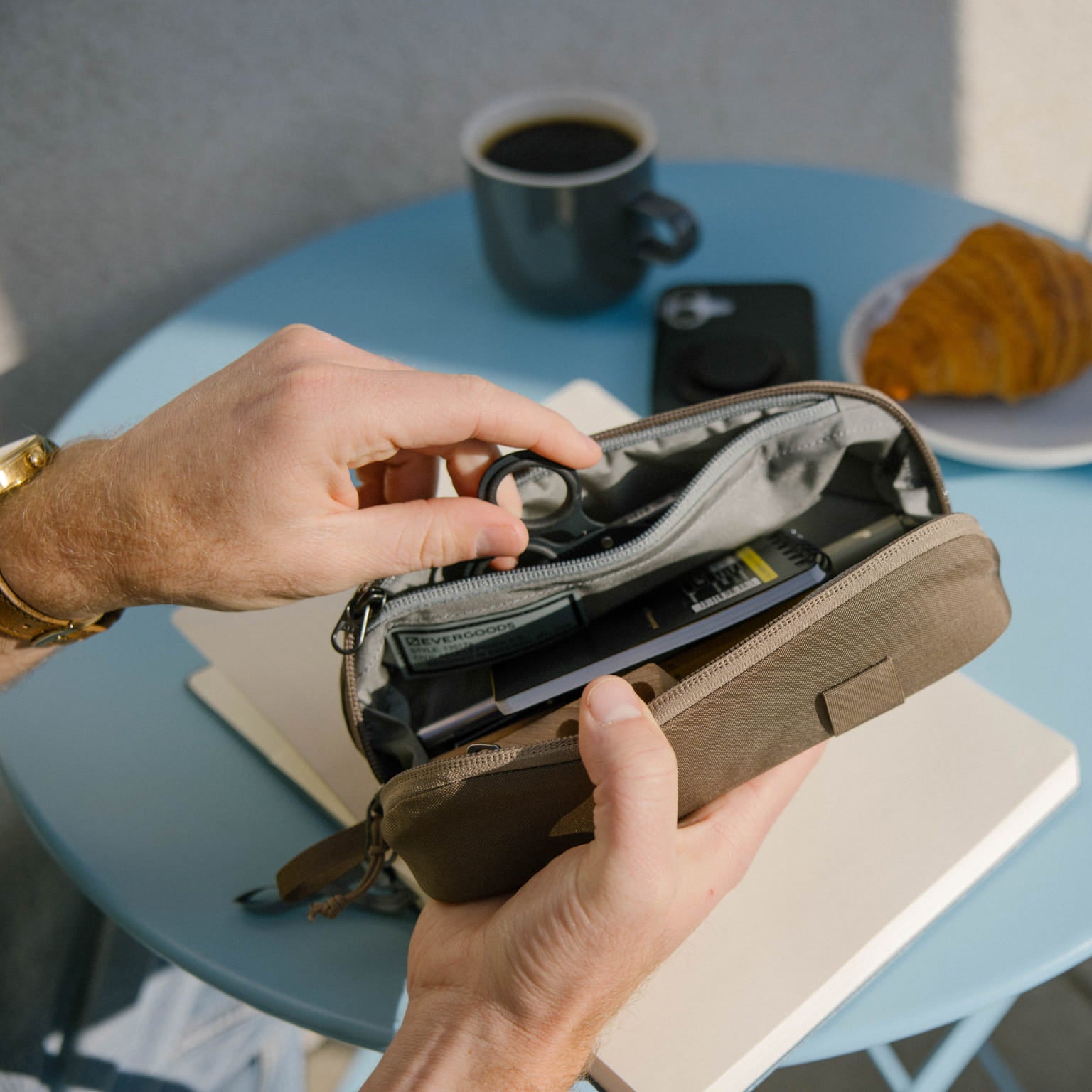 Person opening a CIVIC Access Pouch 0.5L in Fossil Brown on a table with a cup of coffee and croissant in the background [13017a-FBN]