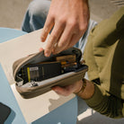 Person opening a CIVIC Access Pouch 0.5L in Fossil Brown travel organizer with items inside, sitting on a blue surface. Person opening a CIVIC Access Pouch 0.5L in Fossil Brown travel organizer with items inside, sitting on a blue surface. [13017a-FBN