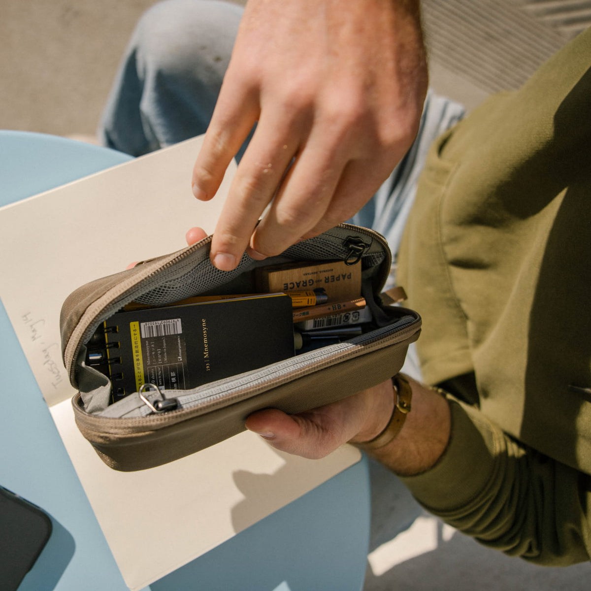 Person opening a CIVIC Access Pouch 0.5L in Fossil Brown travel organizer with items inside, sitting on a blue surface.