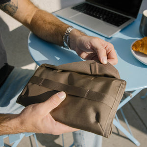 Person holding a CIVIC Access Pouch 1L in Fossil Brown with a laptop and food on a table in the background