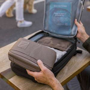 Person packing a CIVIC Access Pouch 2L in Fossil Brown -  travel organizer into an open suitcase on a wooden table.