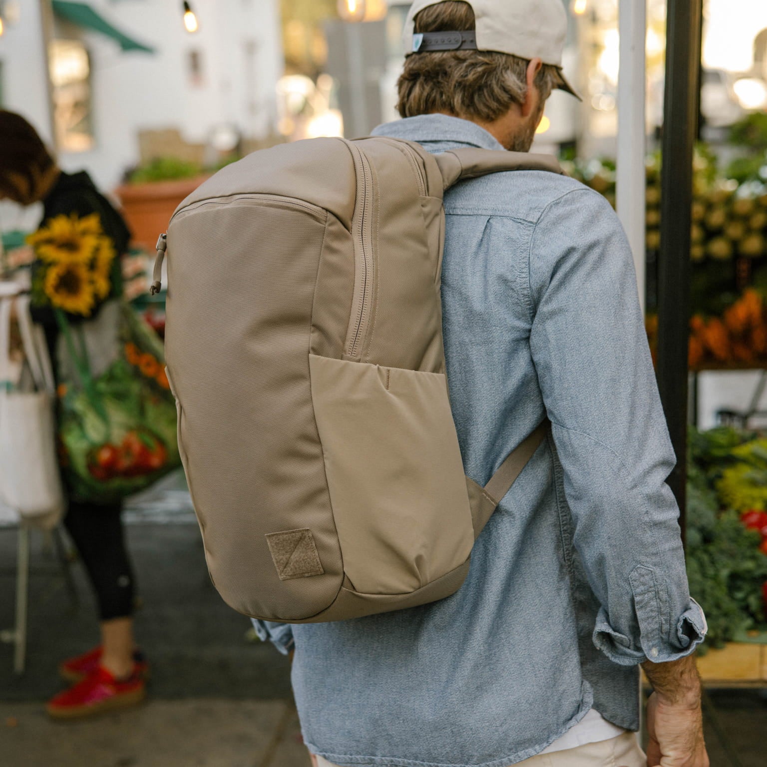 Person wearing a CIVIC Half Zip 22L in Fossil Brown backpack in an outdoor market with people and plants. [13003d-FBN]