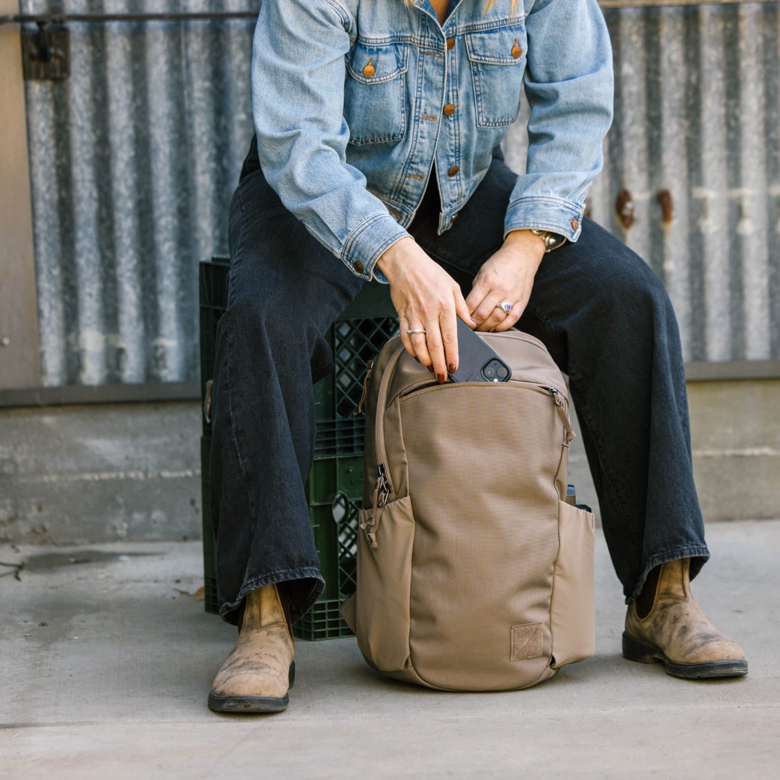 Person sitting on a crate with a CIVIC Half Zip 22L in Fossil Brown backpack, wearing a denim jacket and dark pants. [13003d-FBN]