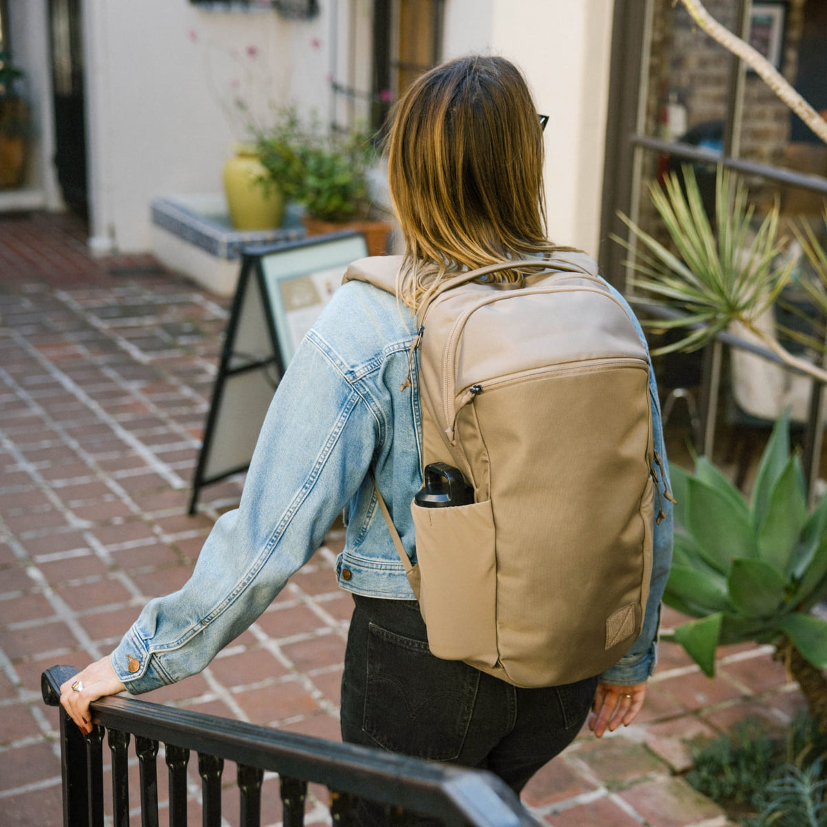 Person wearing a CIVIC Half Zip 22L in Fossil Brown backpack in an outdoor setting with plants and a building in the background