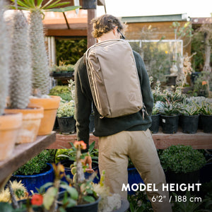 Person with a CIVIC Panel Loader 24L in Fossil Brown  backpack standing in a garden center surrounded by plants.