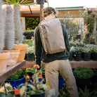 Person with a CIVIC Panel Loader 24L in Fossil Brown  backpack standing in a garden center surrounded by plants. Person with a CIVIC Panel Loader 24L in Fossil Brown  backpack standing in a garden center surrounded by plants. [13001c-FBN