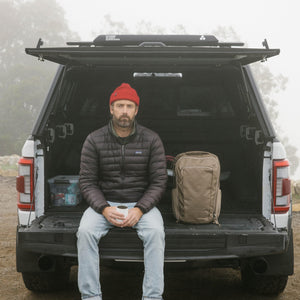 Man sitting in the back of a vehicle with a backpack, wearing a red beanie and dark jacket. CIVIC Travel Bag 20L in Fossil Brown.