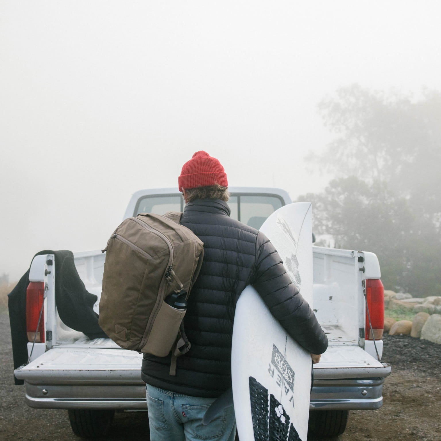 Person with a CIVIC Travel Bag 20L in Fossil Brown and surfboard standing next to a white truck in a foggy landscape. [13019c-FBN]