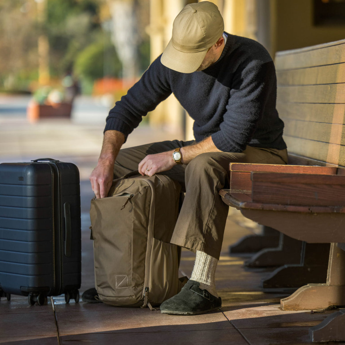 Man sitting on a bench with a CIVIC Travel Bag 26L in Fossil Brown backpack and suitcase accessing front dump quick access pocket.