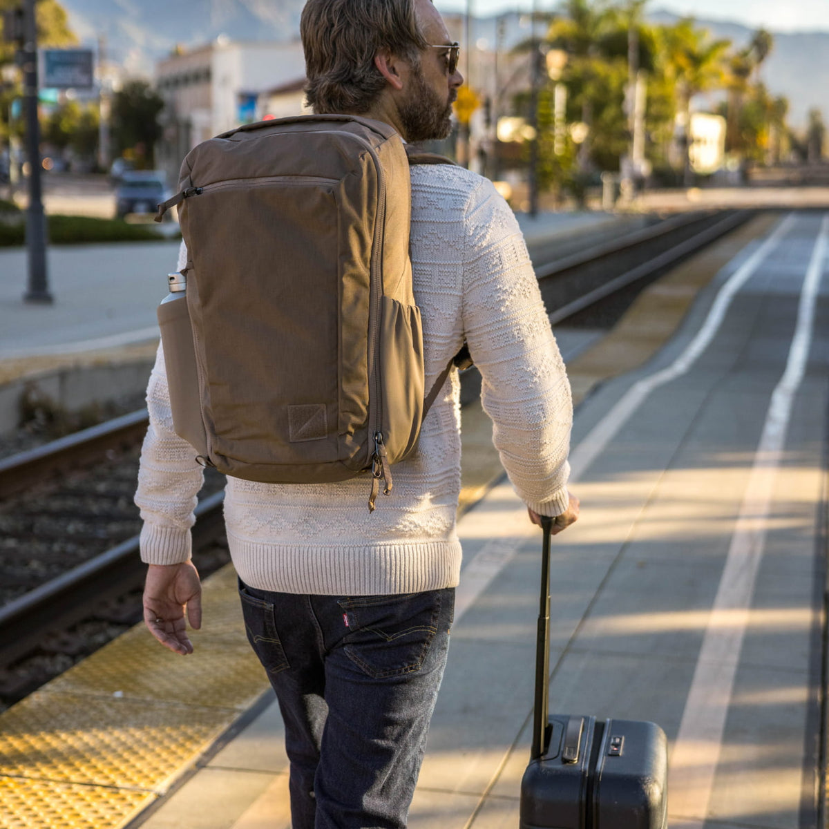 Man with a CIVIC Travel Bag 26L in Fossil Brown backpack and suitcase walking on a train platform