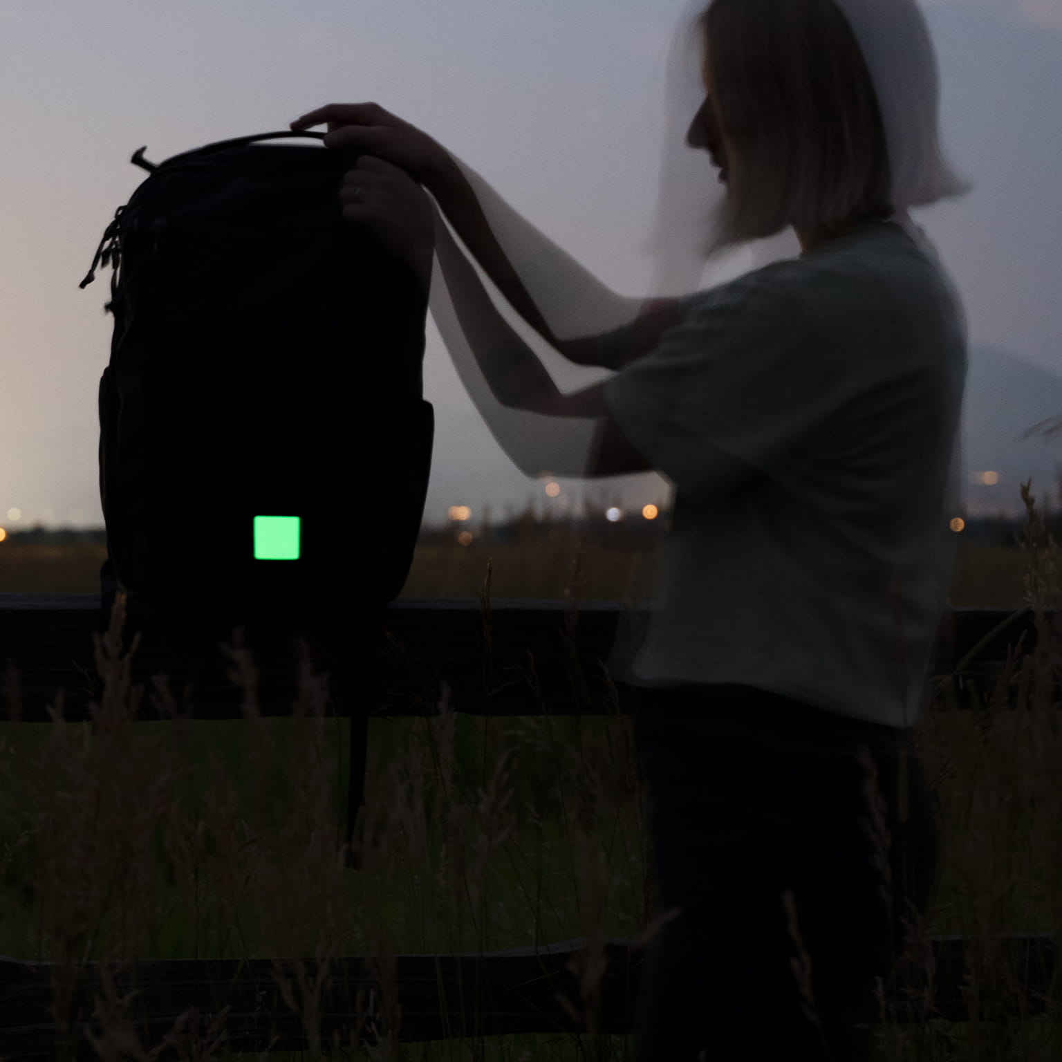 A person outdoors at dusk holds up a black backpack with a glowing green square patch on the front.