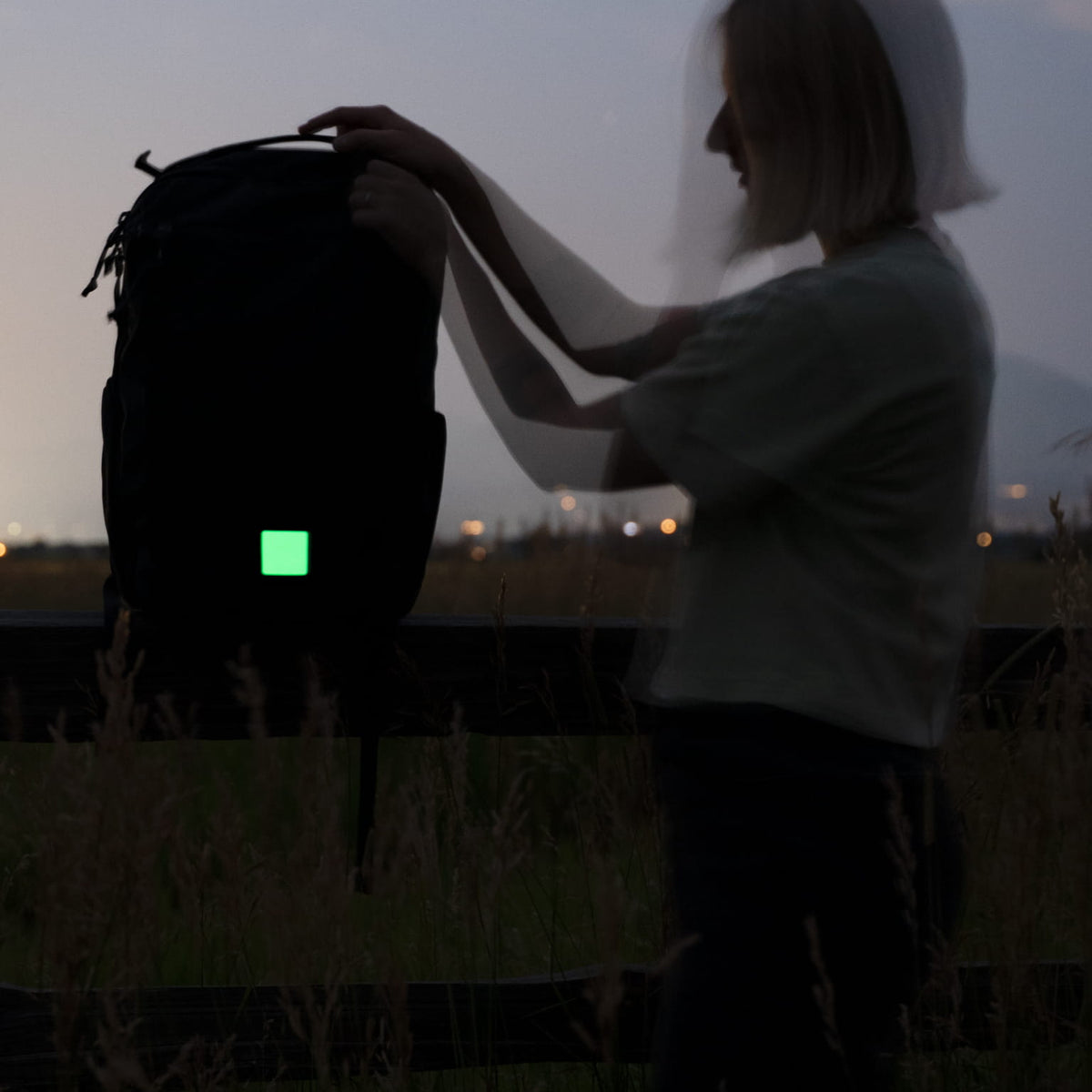 A person outdoors at dusk holds up a black backpack with a glowing green square patch on the front.