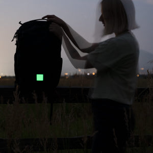A person outdoors at dusk holds up a black backpack with a glowing green square patch on the front.