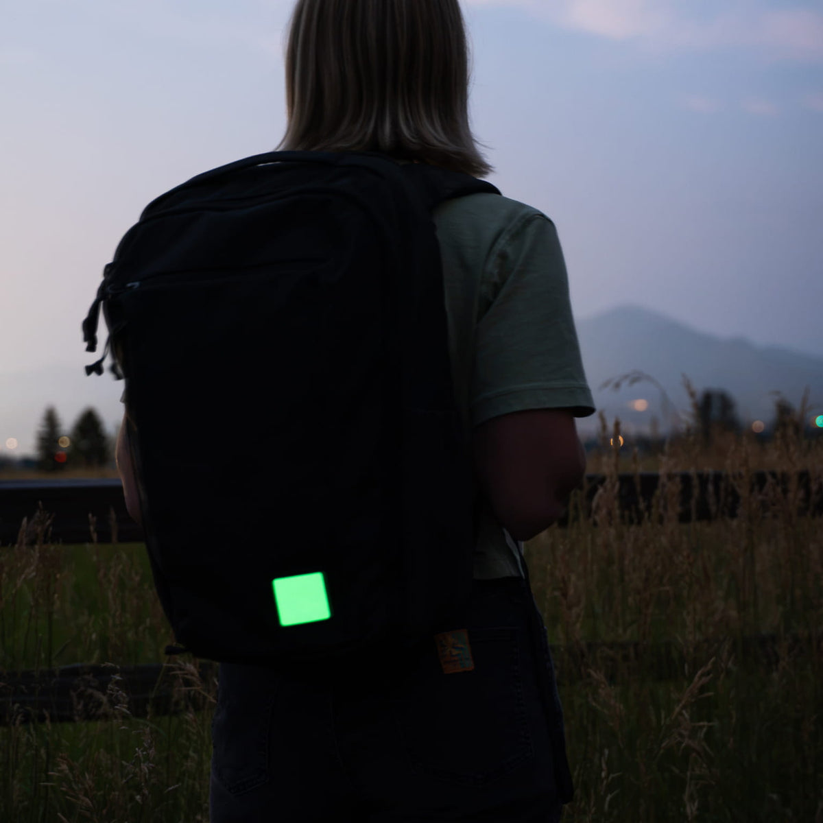 A person viewed from behind wearing a black backpack with a glowing green square patch attached near the bottom, standing outdoors in a field at dusk.