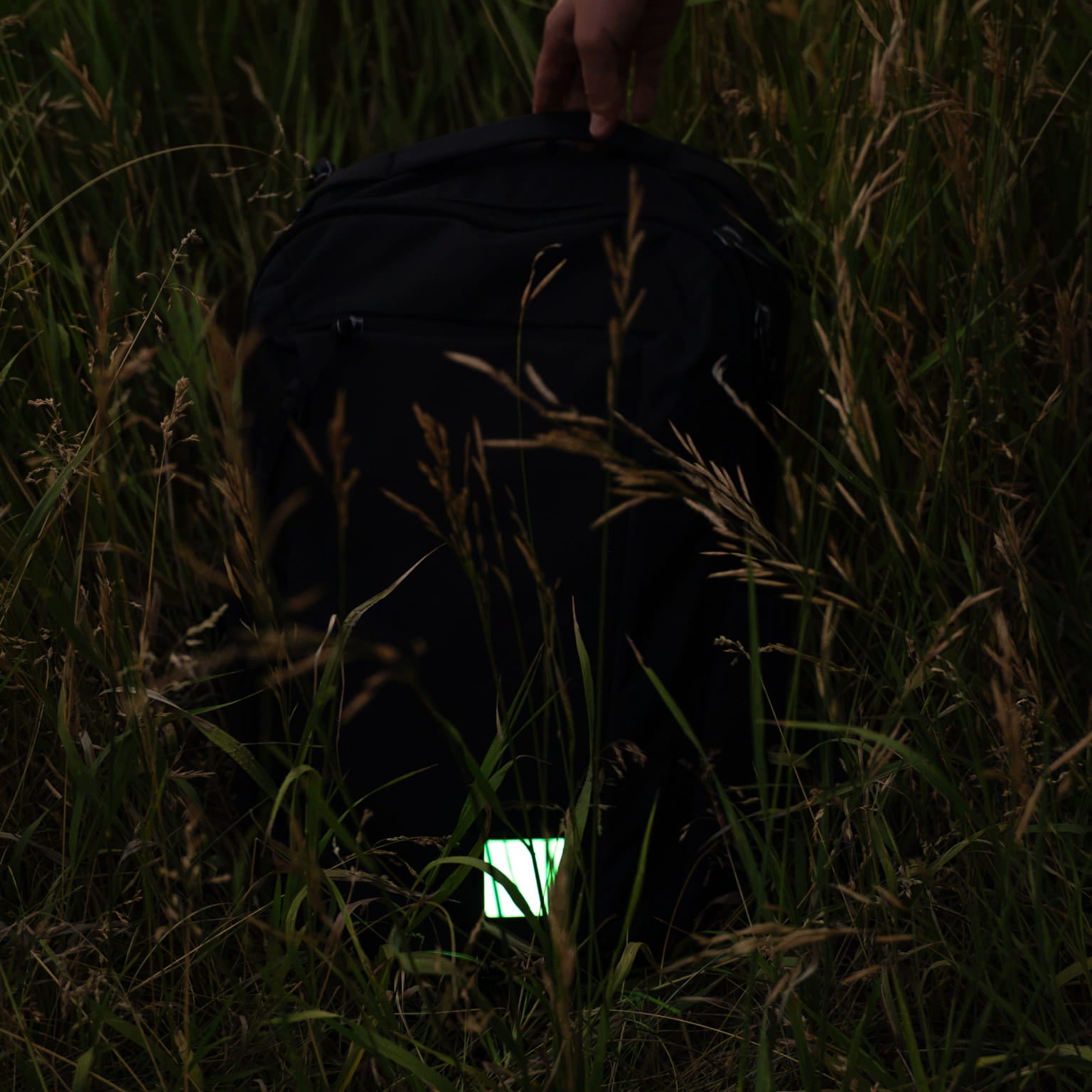 A black backpack lying in tall grass, with a hand holding the top handle and a small glowing patch attached near the bottom front.