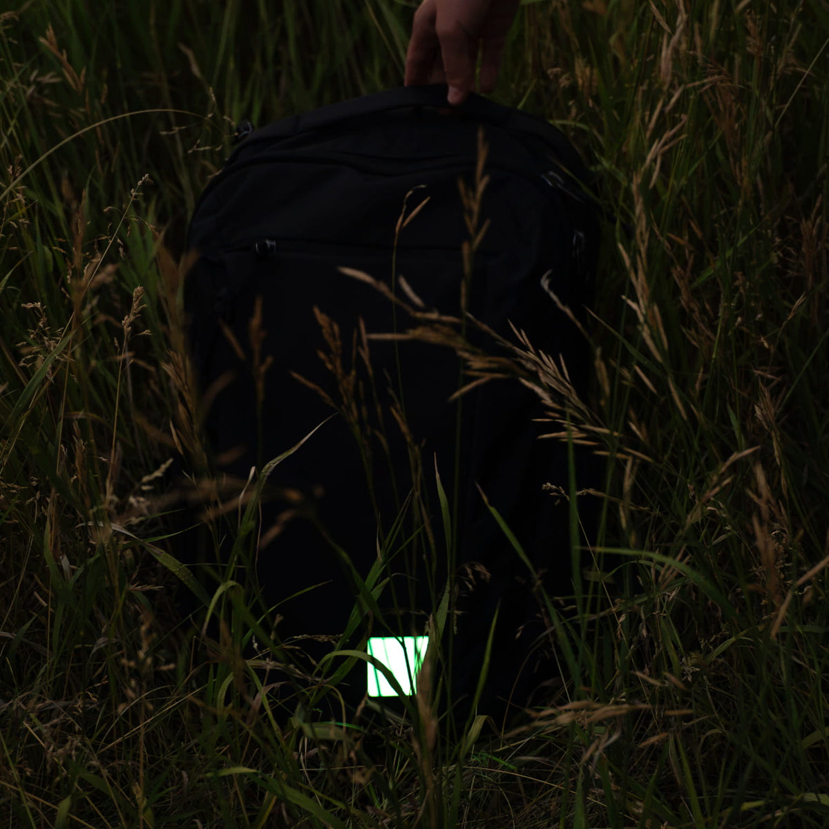A black backpack lying in tall grass, with a hand holding the top handle and a small glowing patch attached near the bottom front.