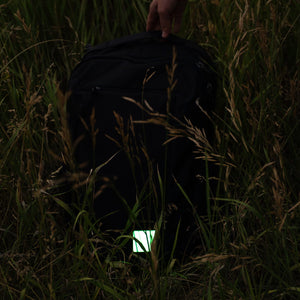 A black backpack lying in tall grass, with a hand holding the top handle and a small glowing patch attached near the bottom front.