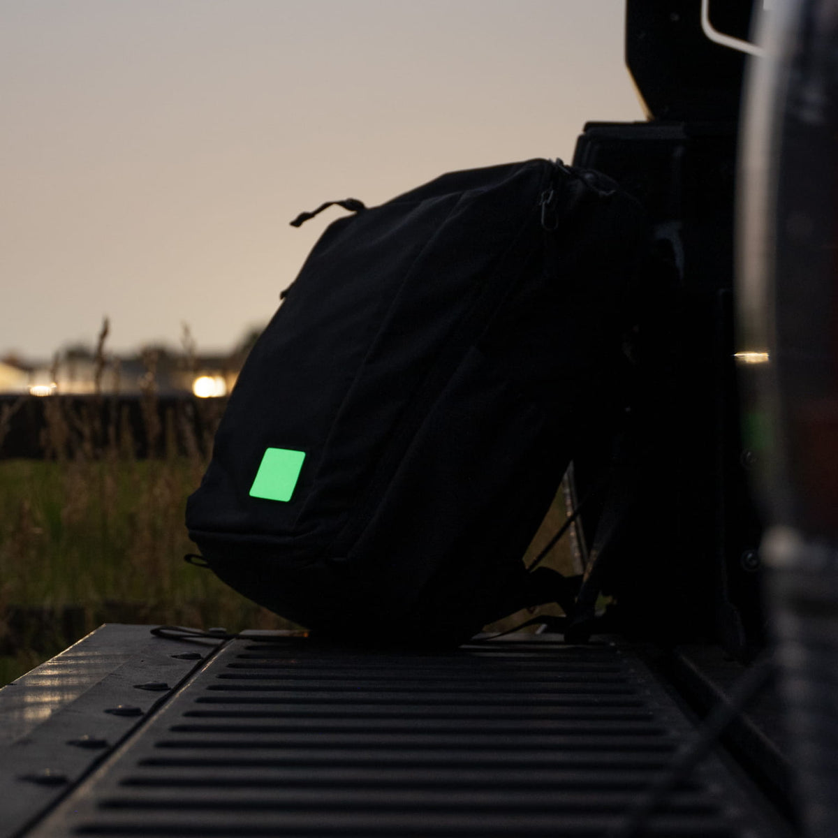 A black backpack with a bright green reflective square patch, resting on the tailgate of a vehicle at dusk.