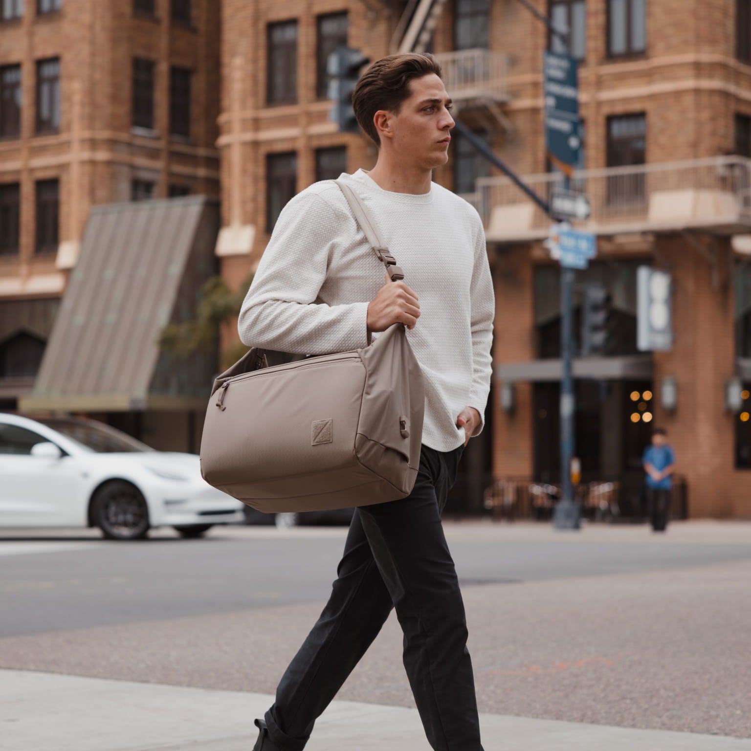 A man in a white sweater and black pants walks across a city street while carrying a large taupe-beige duffel bag over his shoulder. [13023d-FBN]