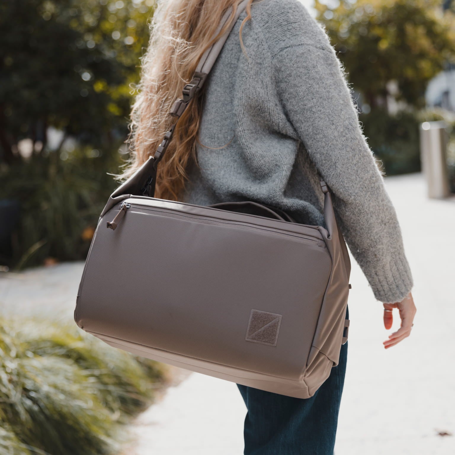 A woman in a gray sweater carries a taupe-gray nylon shoulder bag outdoors on a sunny day. [13023d-FBN]