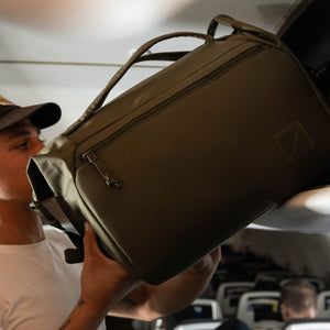 Person pulling a Transit Duffel 25L in OD Green out of an overhead bin inside an airplane