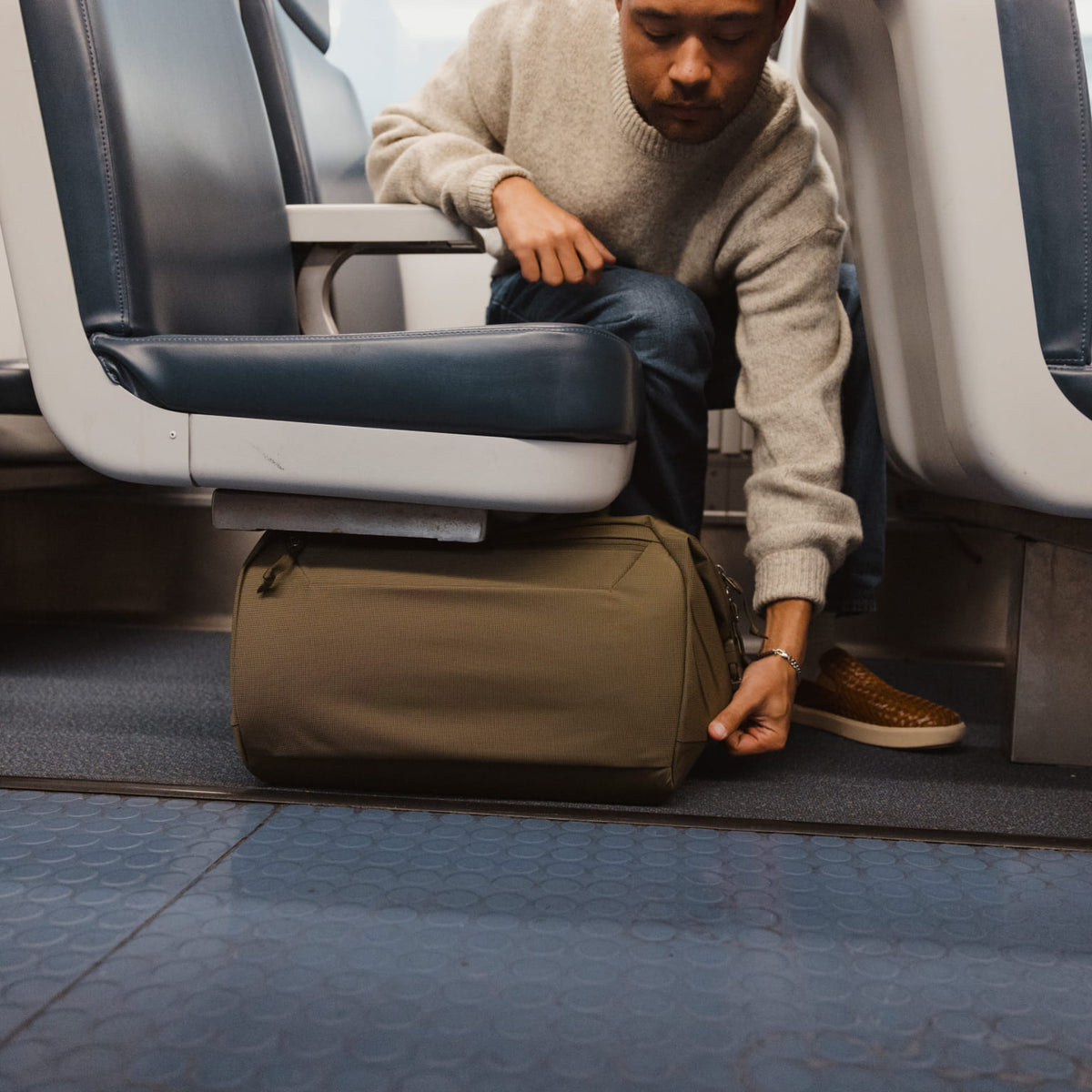 Man adjusting a Transit Duffel 25L in OD Green under an airplane seat