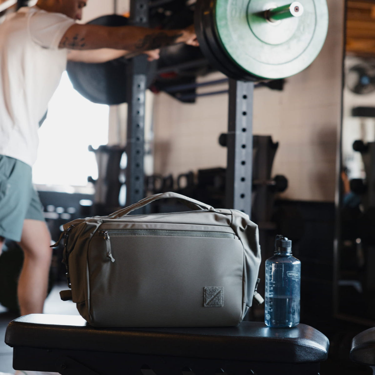 Transit Duffel 25L in OD Green and blue water bottle on a weightlifting bench with a person lifting weights in the background.