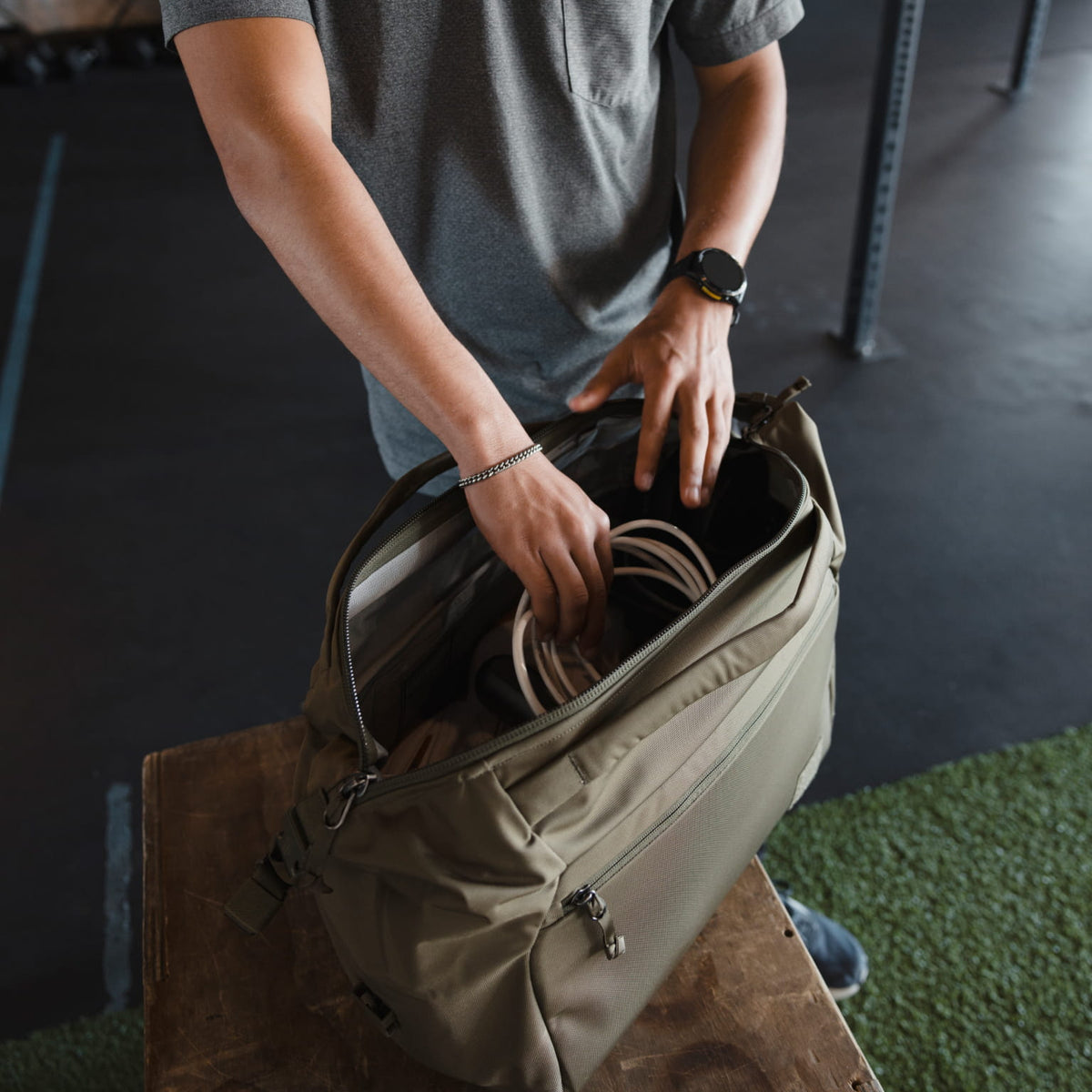 Person organizing items in a Transit Duffel 25L in OD Green on a bench in an indoor gym setting