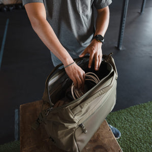 Person organizing items in a Transit Duffel 25L in OD Green on a bench in an indoor gym setting