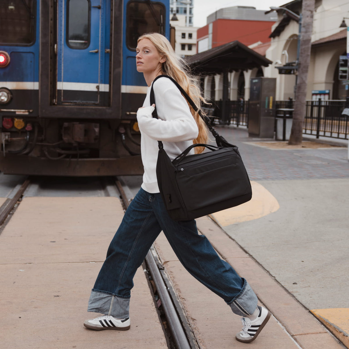 Woman walking past a train with a black duffel, wearing a white sweater and blue jeans.