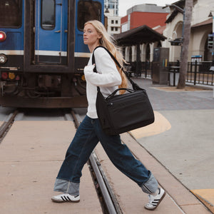 Woman walking past a train with a black duffel, wearing a white sweater and blue jeans.